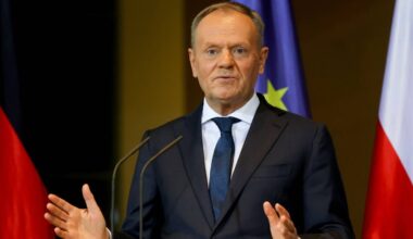 Donald Tusk, polityk w ciemnym garniturze i niebieskim krawacie, przemawia za mównicą. Za nim widoczne są flagi Polski i Niemiec.