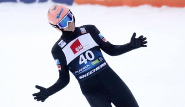 Skoczek narciarski w czarnym kombinezonie z numerem 40 na piersi, w r&oacute;żowym kasku i niebieskich goglach, z rękami rozłożonymi na boki.