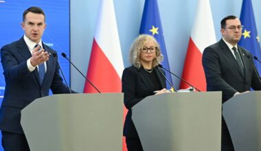Trzy osoby stoją za m&oacute;wnicami na tle flag Polski i Unii Europejskiej. Mężczyzna po lewej gestykuluje ręką.