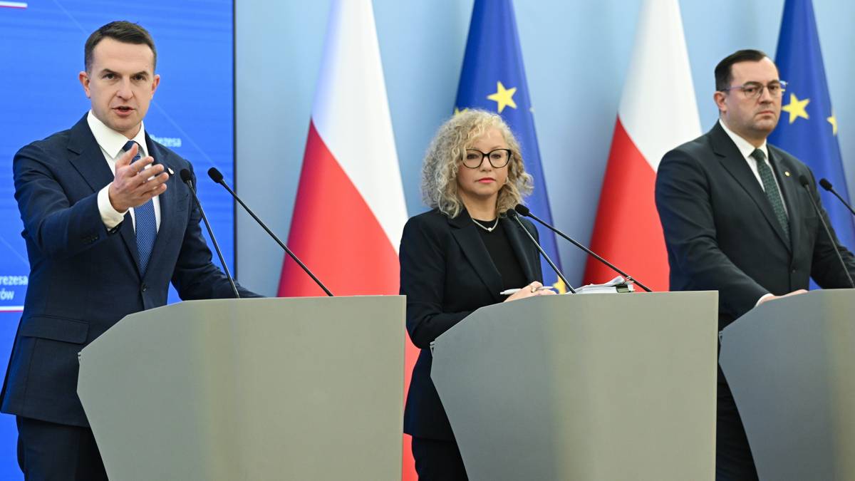 Trzy osoby stoją za m&oacute;wnicami na tle flag Polski i Unii Europejskiej. Mężczyzna po lewej gestykuluje ręką.