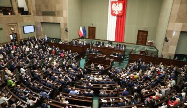 Widok z góry na salę plenarną Sejmu Rzeczypospolitej Polskiej, z posłami siedzącymi w ławach i flagą Polski na ścianie.