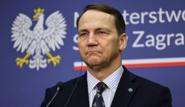 Radosław Sikorski, polski minister spraw zagranicznych, stoi przed niebieskim tłem z godłem Polski.