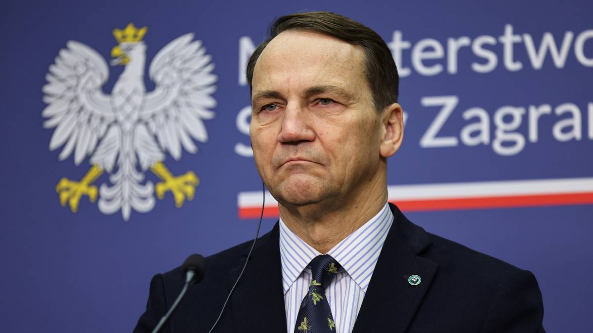 Radosław Sikorski, polski minister spraw zagranicznych, stoi przed niebieskim tłem z godłem Polski.