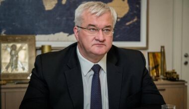 Mężczyzna w garniturze i krawacie siedzi przy biurku, za nim widoczna jest mapa świata.