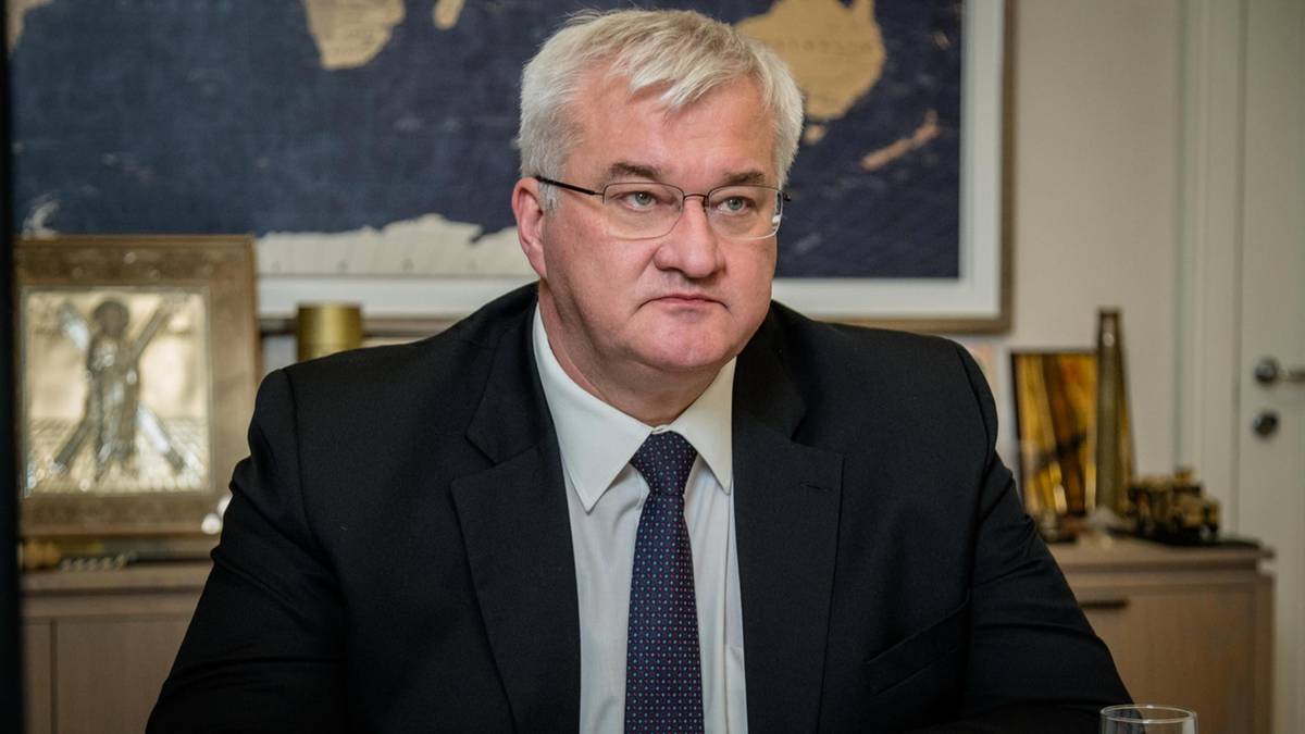 Mężczyzna w garniturze i krawacie siedzi przy biurku, za nim widoczna jest mapa świata.