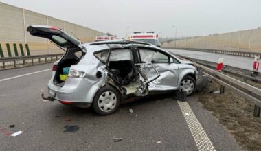 Srebrny samoch&oacute;d osobowy po wypadku na autostradzie, z otwartymi drzwiami i widocznymi uszkodzeniami karoserii. Na drodze leżą fragmenty samochodu i znaki.