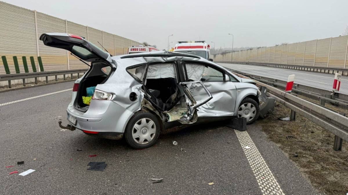 Srebrny samoch&oacute;d osobowy po wypadku na autostradzie, z otwartymi drzwiami i widocznymi uszkodzeniami karoserii. Na drodze leżą fragmenty samochodu i znaki.