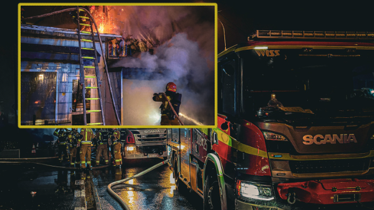 Strażacy gaszą pożar na targowisku, widać płonące kontenery morskie i dym unoszący się w powietrzu.