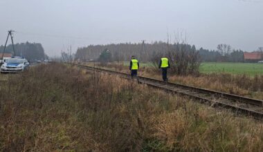 Dwóch policjantów w żółtych kamizelkach idzie wzdłuż torów kolejowych. W tle widać samochód policyjny i drzewa.