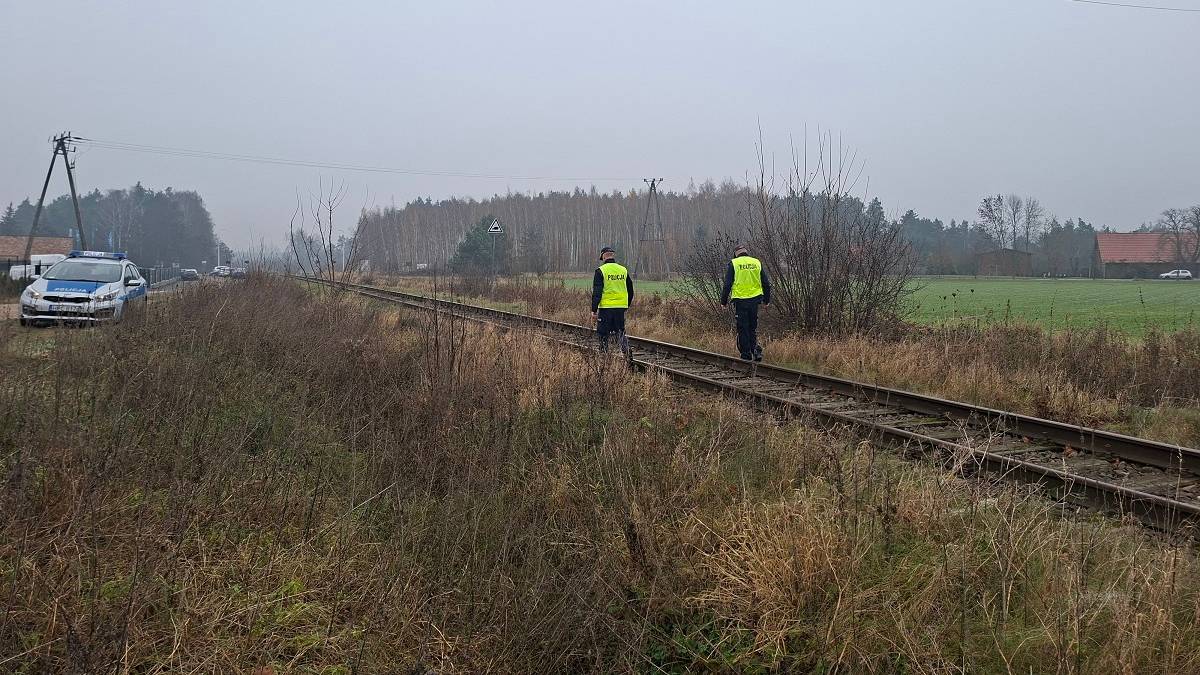 Dwóch policjantów w żółtych kamizelkach idzie wzdłuż torów kolejowych. W tle widać samochód policyjny i drzewa.