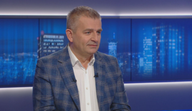 Mężczyzna w białej koszuli i marynarce w kratę siedzi w studiu telewizyjnym.