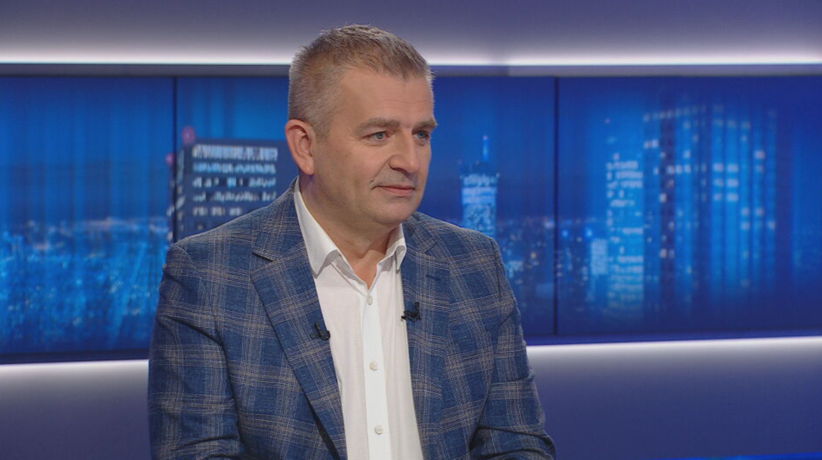 Mężczyzna w białej koszuli i marynarce w kratę siedzi w studiu telewizyjnym.