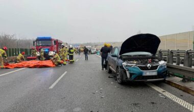 Służby ratunkowe działają na miejscu wypadku drogowego na autostradzie. Widać samoch&oacute;d osobowy z otwartą maską, stojący na poboczu, oraz strażak&oacute;w zajmujących się osobą na kocu ratunkowym na pasie ruchu. W tle stoi ciężar&oacute;wka.