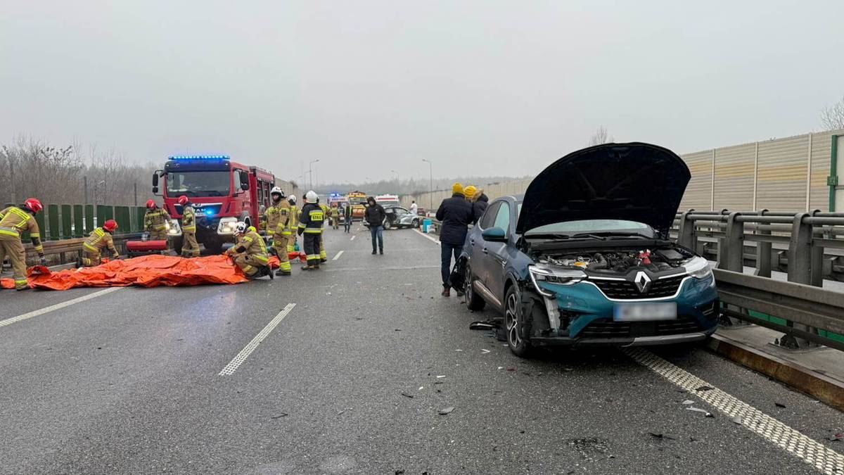 Służby ratunkowe działają na miejscu wypadku drogowego na autostradzie. Widać samoch&oacute;d osobowy z otwartą maską, stojący na poboczu, oraz strażak&oacute;w zajmujących się osobą na kocu ratunkowym na pasie ruchu. W tle stoi ciężar&oacute;wka.