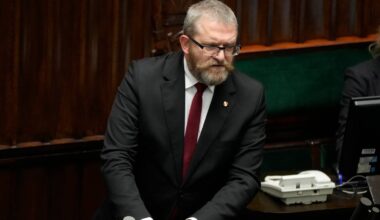 Grzegorz Braun w garniturze i krawacie stoi przy m&oacute;wnicy w sali plenarnej, opierając ręce na pulpicie.