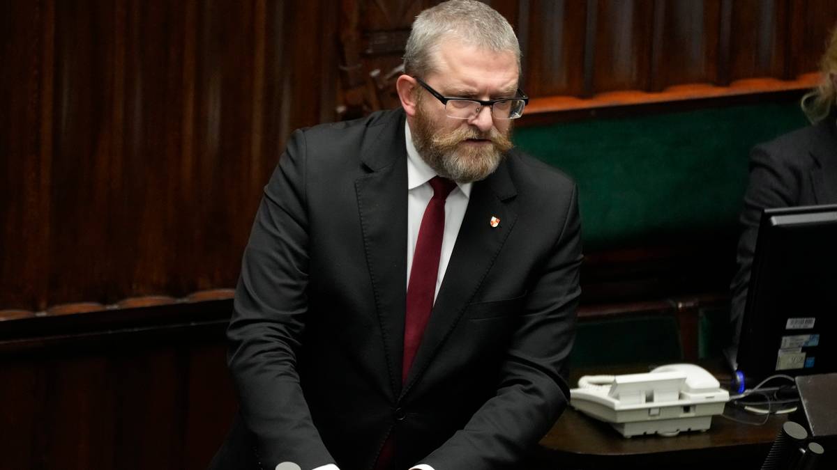 Grzegorz Braun w garniturze i krawacie stoi przy m&oacute;wnicy w sali plenarnej, opierając ręce na pulpicie.