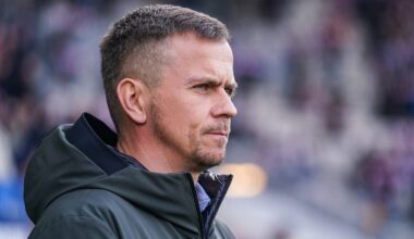 Portret trenera piłkarskiego w zbliżeniu, w ciemnej kurtce, na tle stadionu.