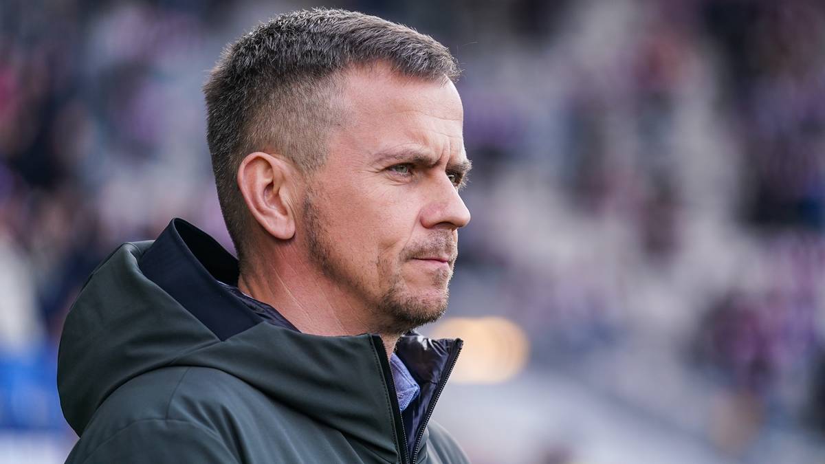 Portret trenera piłkarskiego w zbliżeniu, w ciemnej kurtce, na tle stadionu.