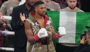 Pięściarz Anthony Joshua w rękawicach bokserskich, ubrany w kamizelkę z motywem kamuflażu, stoi przed flagą Nigerii.