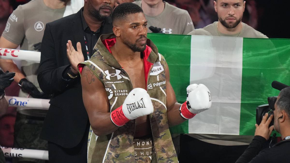 Pięściarz Anthony Joshua w rękawicach bokserskich, ubrany w kamizelkę z motywem kamuflażu, stoi przed flagą Nigerii.