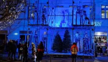 Budynek oświetlony jaskrawym, niebieskim światłem. Na fasadzie widoczne są rusztowania i podświetlone figury, przypominające postacie z bajek lub scenografię teatralną. Przed budynkiem stoi grupa ludzi, obserwujących ekspozycję.