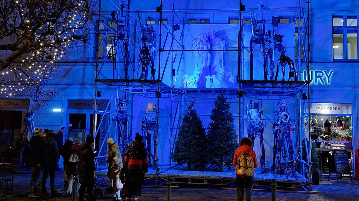 Budynek oświetlony jaskrawym, niebieskim światłem. Na fasadzie widoczne są rusztowania i podświetlone figury, przypominające postacie z bajek lub scenografię teatralną. Przed budynkiem stoi grupa ludzi, obserwujących ekspozycję.