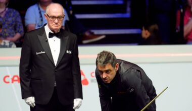 Sędzia snookerowy w czarnym fraku i białych rękawiczkach stoi obok zawodnika w czarnej koszulce, kt&oacute;ry przygotowuje się do uderzenia kijem w bilę na zielonym stole.