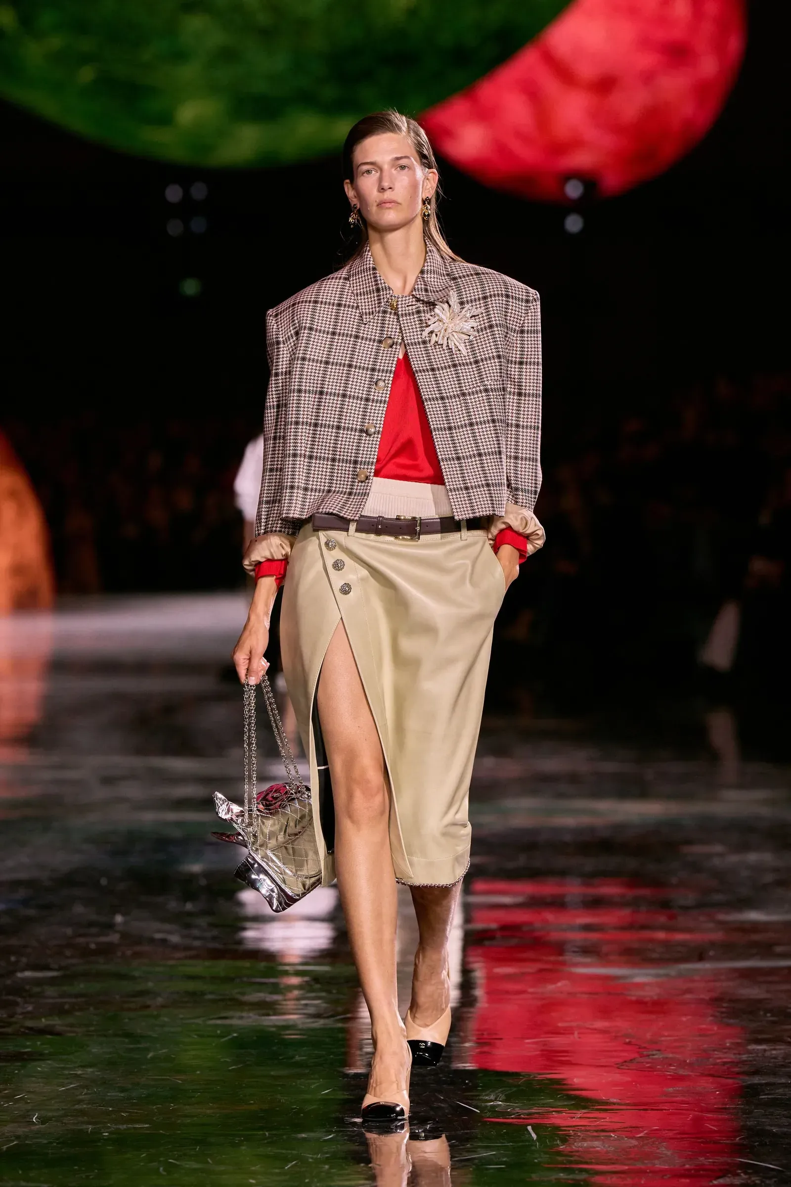 Modelka na wybiegu w stylowej, kraciastej kurtce i czerwonym topie, z ołówkową spódnicą i eleganckimi dodatkami.