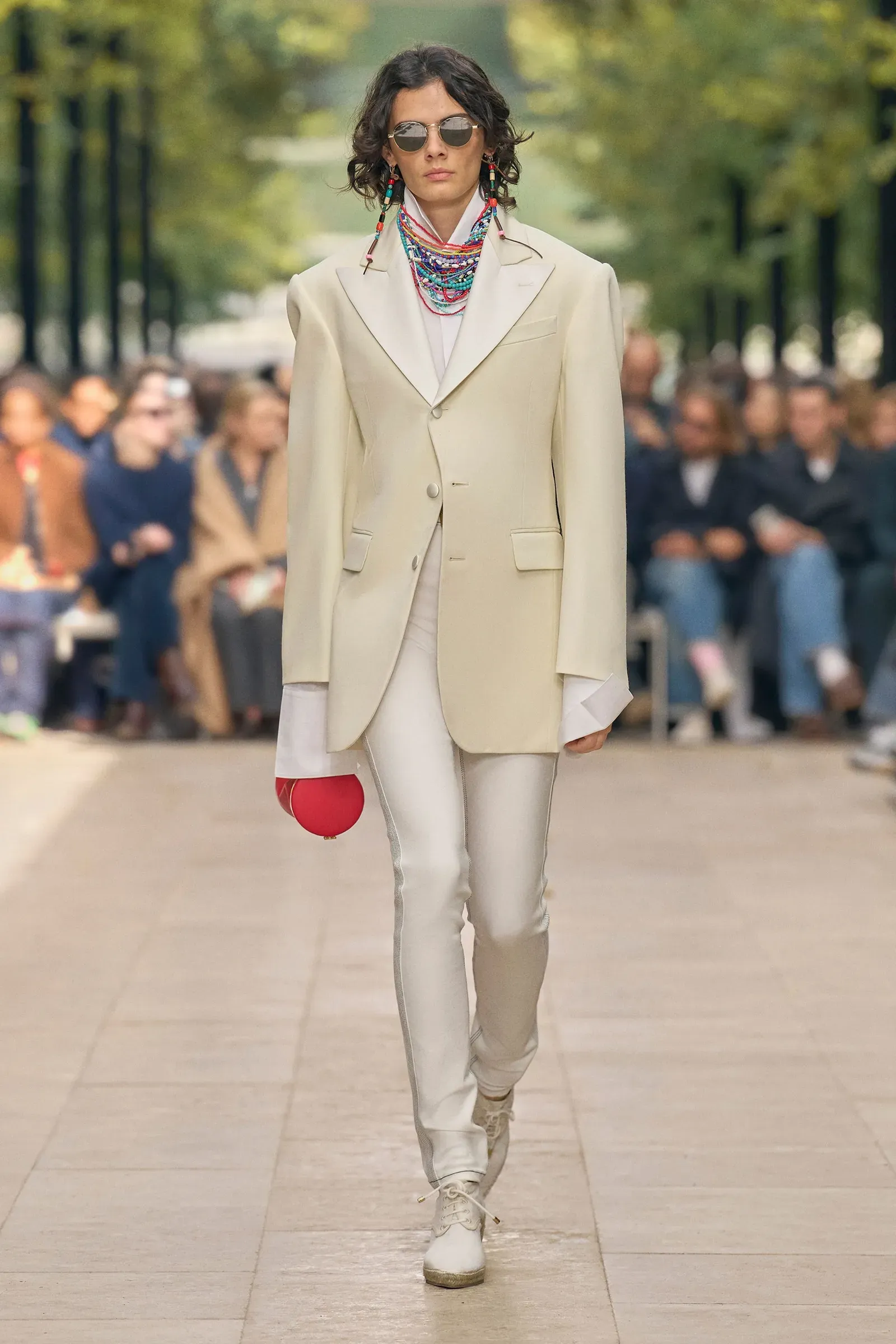 Modelka w jasnym garniturze i okularach przeciwsłonecznych, idzie po wybiegu, trzymając czerwoną piłkę. W tle widownia.