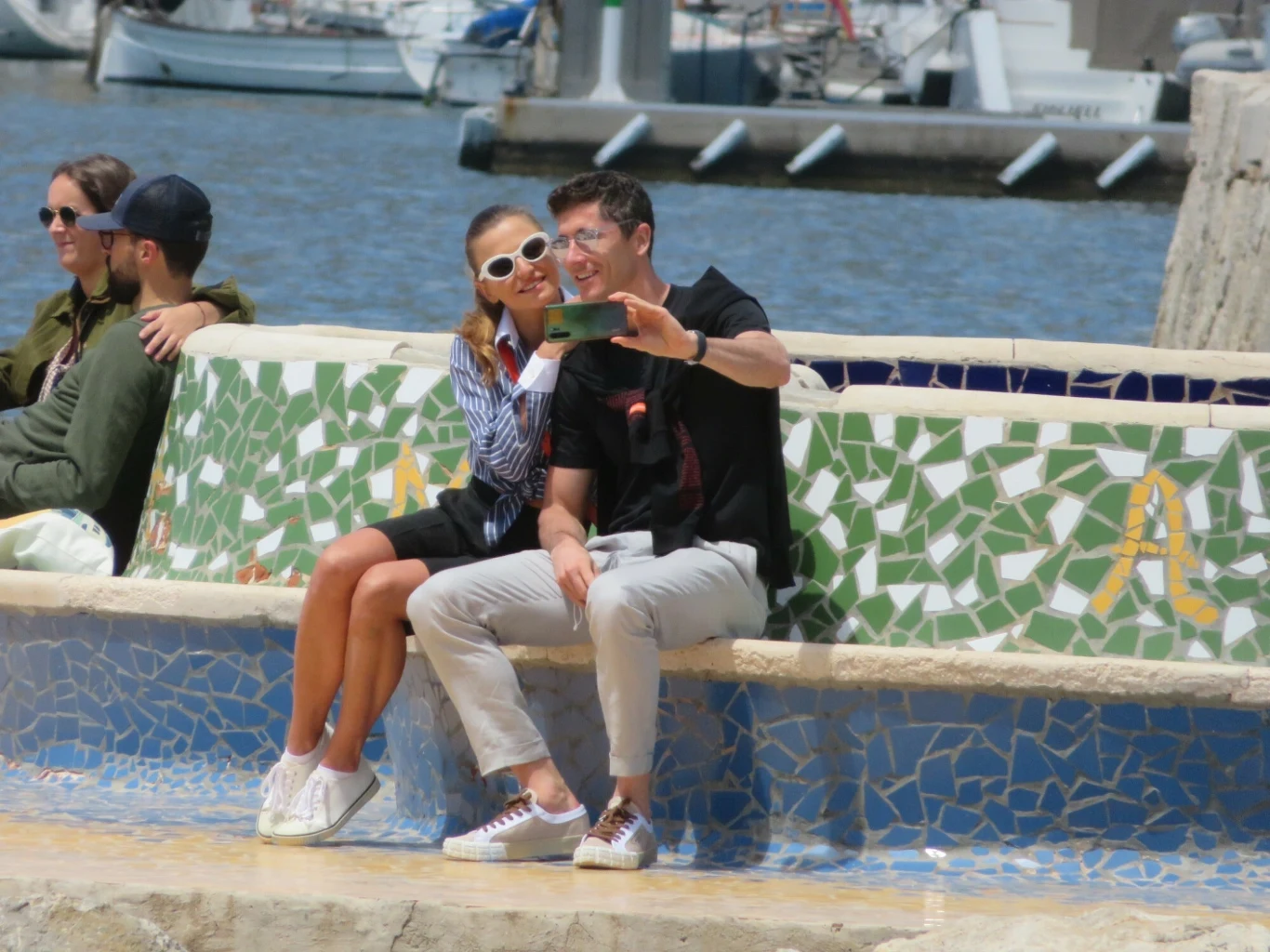 Para robiąca sobie selfie na kolorowej mozaikowej ławce przy marinie, w tle widać wodę i zacumowane łodzie, obok nich siedzi inna para, wszyscy sprawiają wrażenie zrelaksowanych i pogodnych.