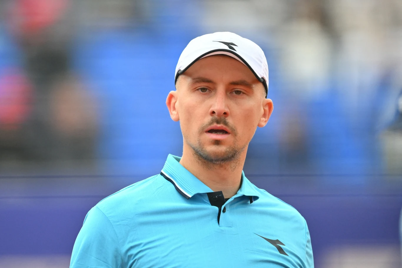 Mężczyzna w sportowej czapce i błękitnej koszulce polo stoi na tle rozmytych trybun stadionu, patrząc poważnie przed siebie.