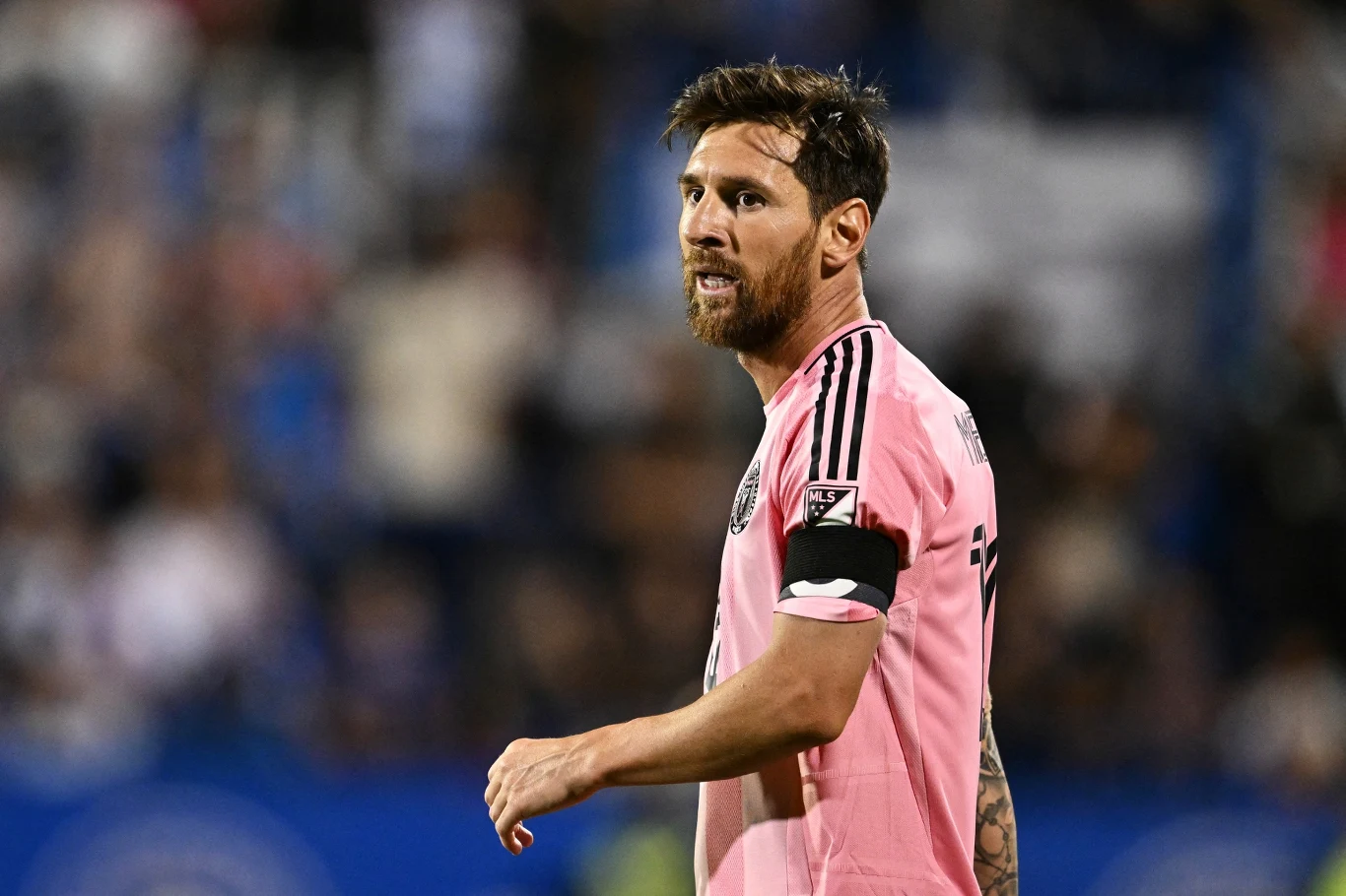 Piłkarz w różowej koszulce z opaską kapitana na ramieniu, w tle rozmyta publiczność i stadionowe światła.