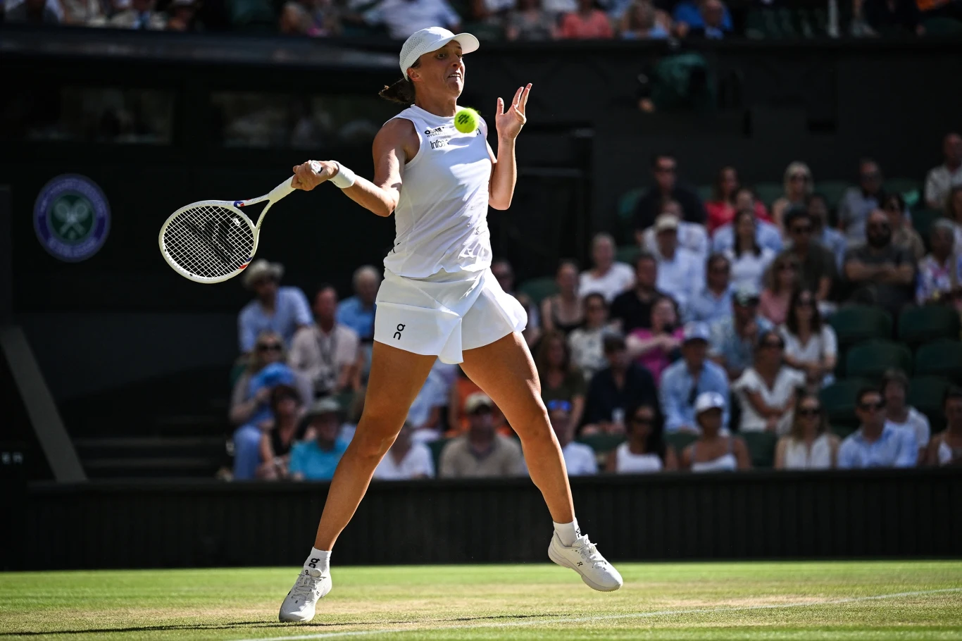 Iga Świątek w trakcie Wimbledonu 2025 Tenisistka w białym stroju wykonuje dynamiczne uderzenie na korcie trawiastym, w tle widownia oraz elementy stadionu.