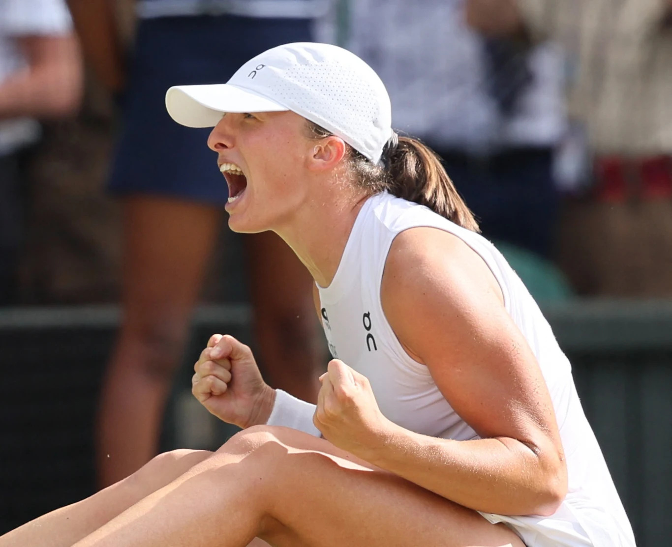 Iga Świątek wygrała Wimbledon 2025 Tenisistka w białym stroju i czapce zaciska pięści, wyraża silne emocje i radość po udanym zagraniu, siedząc na korcie tenisowym.