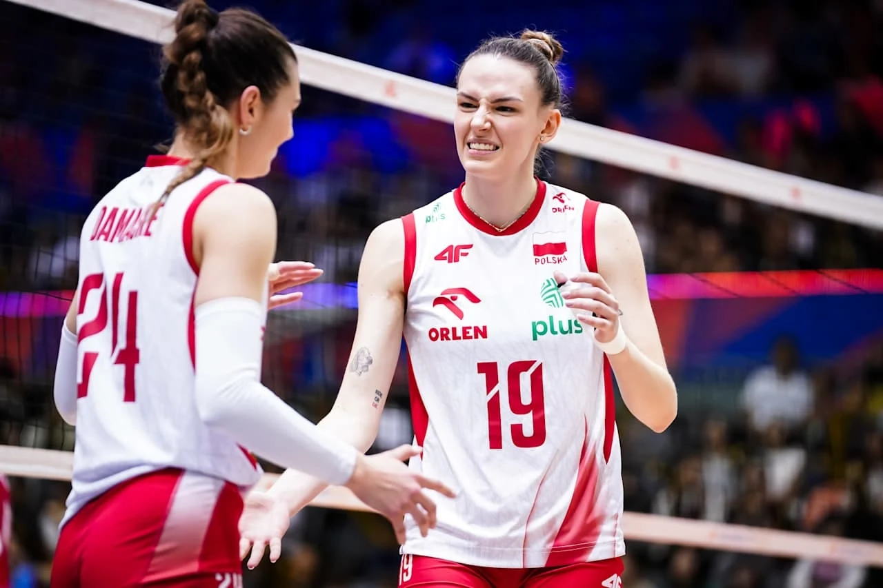 Dwie siatkarki reprezentacji Polski w trakcie meczu, ubrane w oficjalne stroje sportowe, wymieniające się gestami i emocjami na boisku, w tle widoczne siatka i rozmyta publiczność.