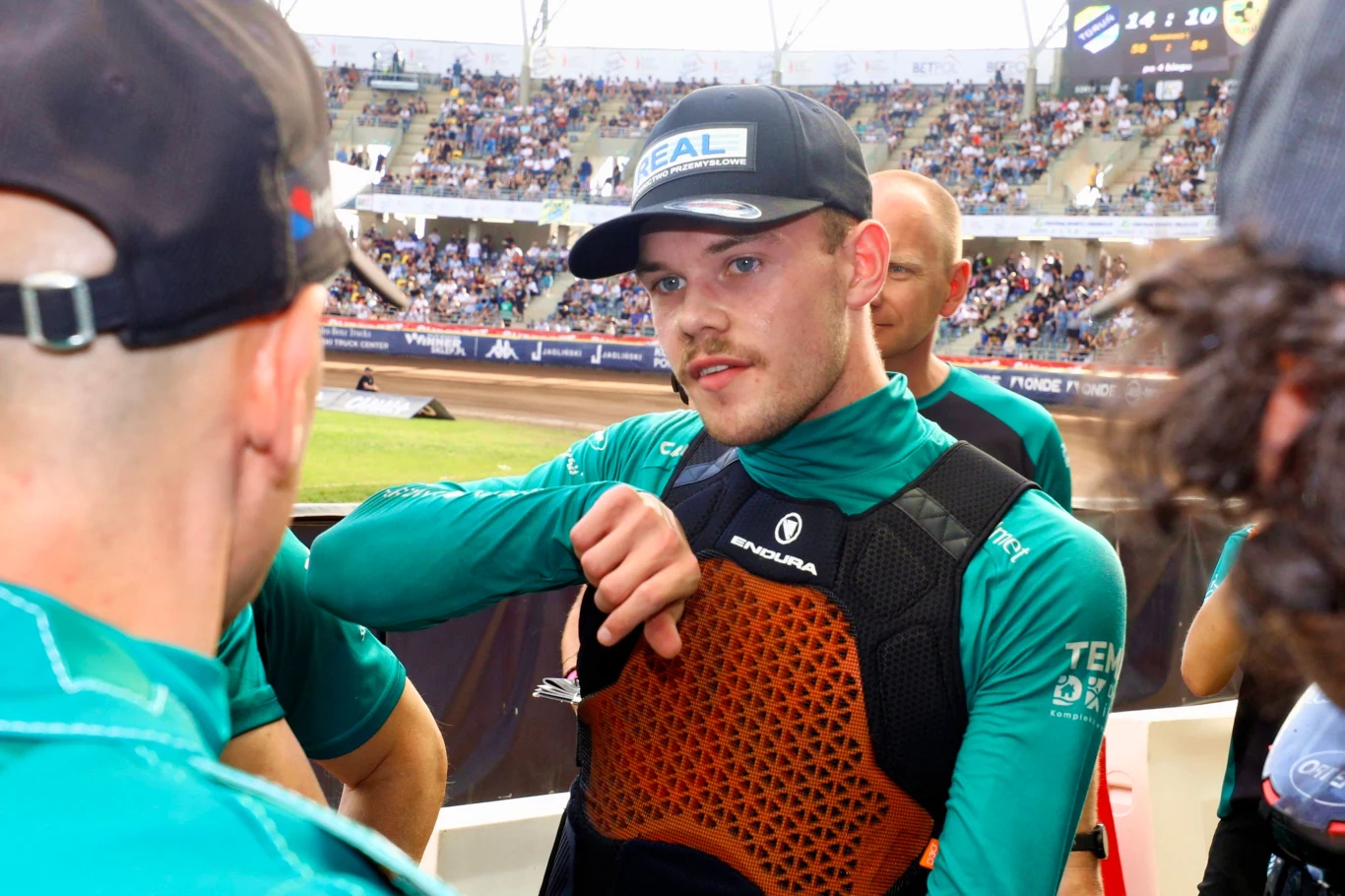 Młody mężczyzna w sportowym stroju i czapce z daszkiem, otoczony przez innych członków drużyny, stoi na stadionie żużlowym podczas zawodów, widząc za plecami trybuny pełne kibiców.