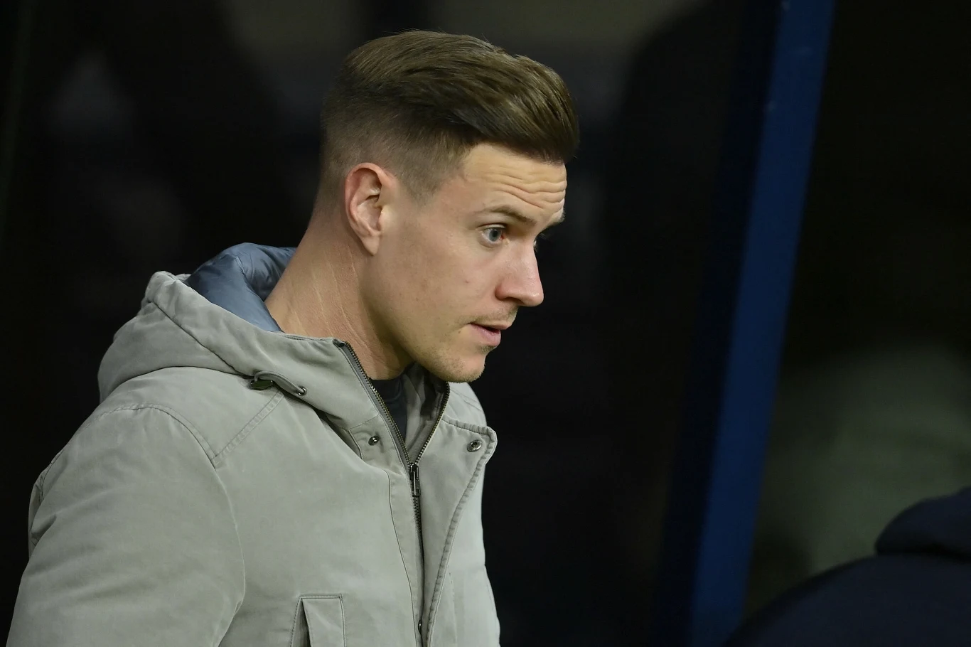 Mężczyzna w szarej kurtce z krótkimi, starannie ułożonymi włosami patrzy w bok, stoi w pobliżu ciemnego tła.