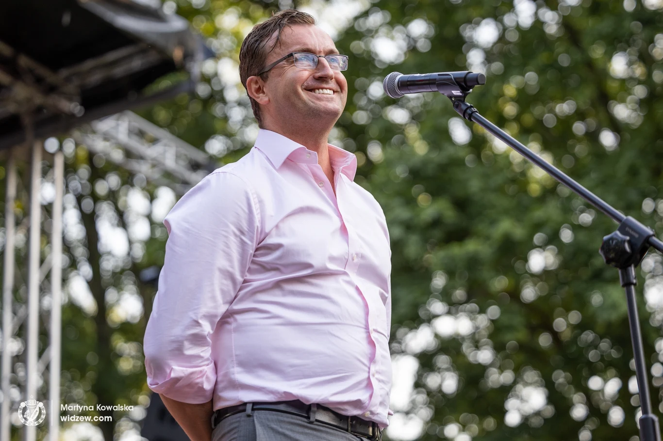 Uśmiechnięty mężczyzna w okularach i różowej koszuli stoi przy mikrofonie na scenie plenerowej, w tle widoczne rozmyte liście drzew.