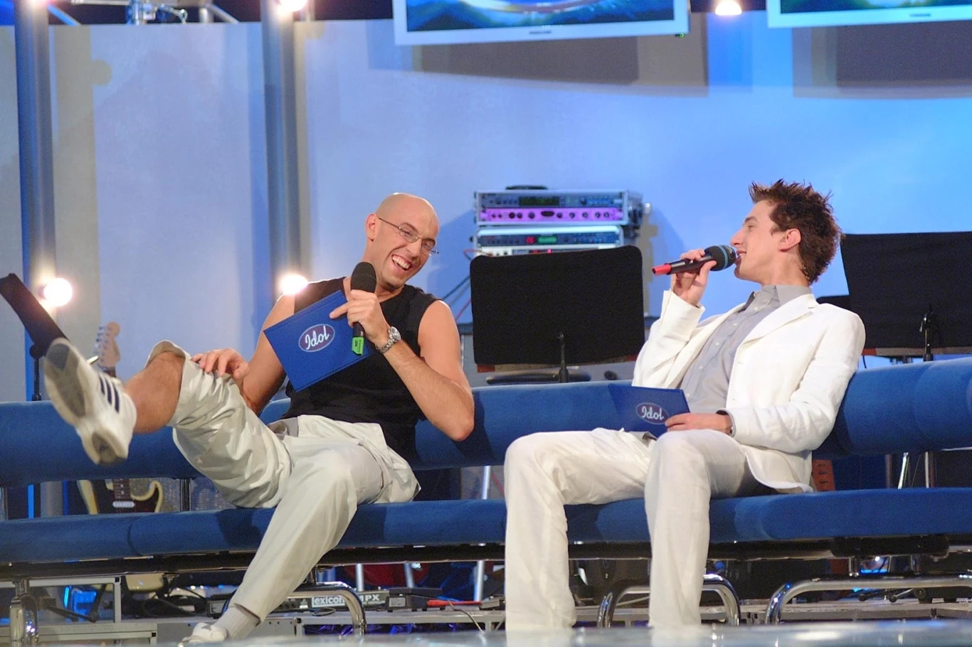 Dwóch mężczyzn siedzi na niebieskiej kanapie w studiu telewizyjnym, trzymają mikrofony i rozmawiają ze sobą, w tle widoczne są elementy wyposażenia scenicznego oraz ekrany.