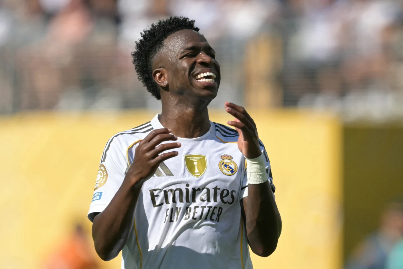 Zawodnik w białym stroju klubu Real Madryt cieszy się podczas meczu piłki nożnej, wyrażając radość z gry na boisku, w tle nieostre postacie kibiców i żółta ściana stadionu