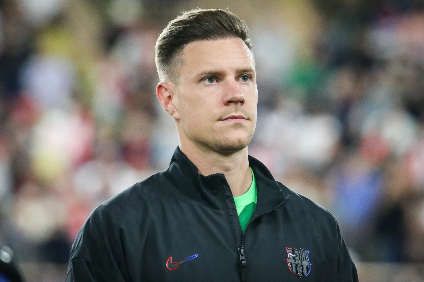 Mężczyzna w sportowej kurtce z herbem klubu FC Barcelona, patrzący przed siebie, tło rozmyte z obecnością publiczności.