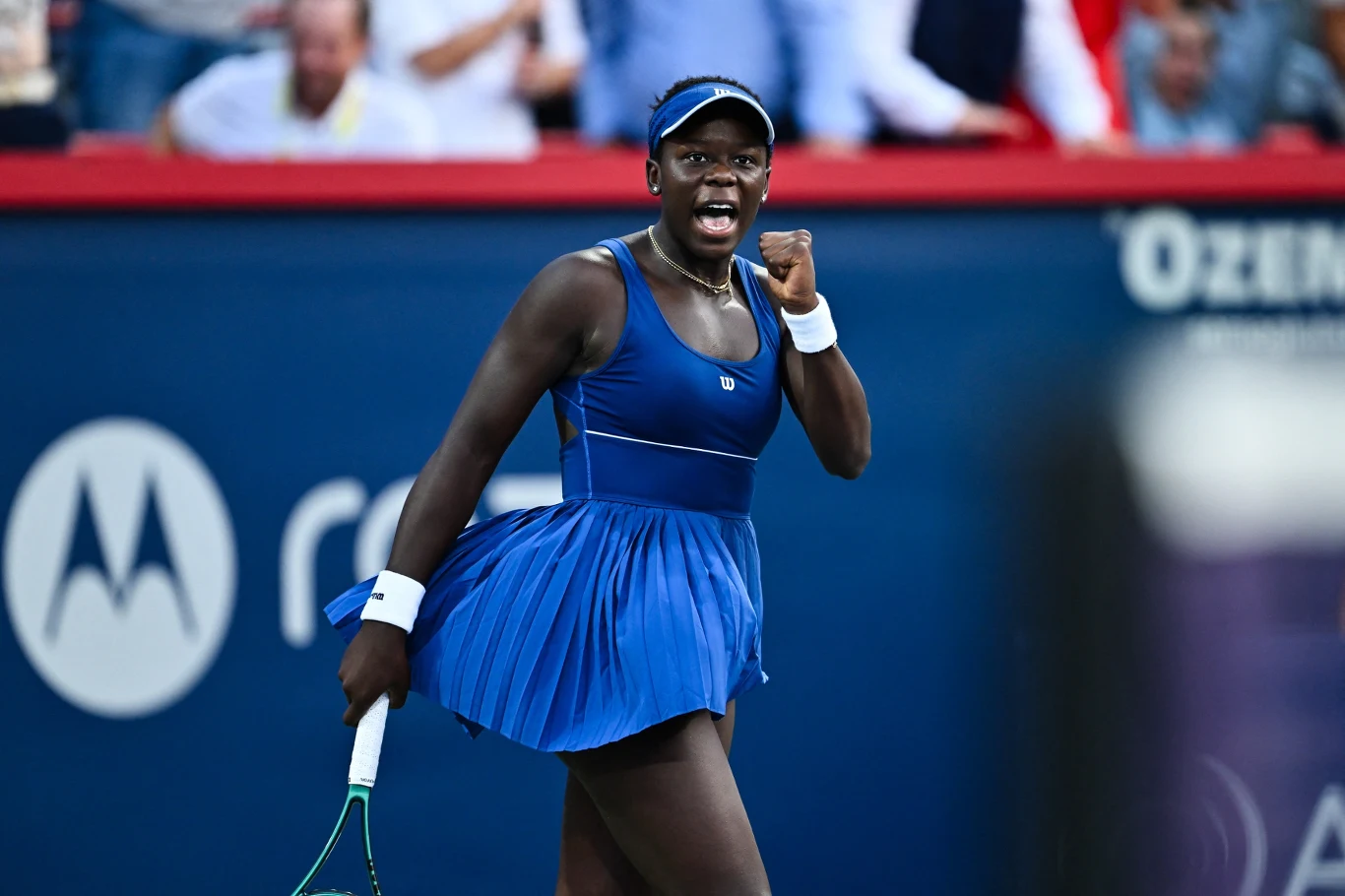 Zawodniczka tenisowa ubrana w niebieską sukienkę sportową po udanym punkcie na korcie, dynamicznie zaciśnięta pięść i wyraźna emocja na twarzy, w tle rozmazana publiczność podczas turnieju.