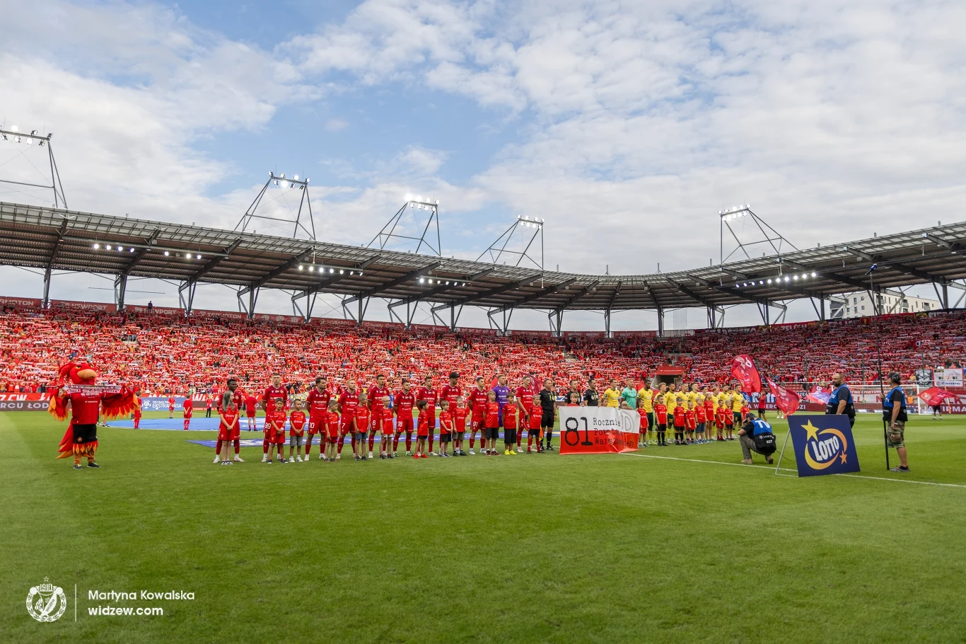 Piłkarze dwóch drużyn ustawieni w rzędzie na murawie stadionu przed rozpoczęciem meczu, trybuny wypełnione kibicami ubranymi głównie na czerwono, widoczne flagi i maskotki drużyn.