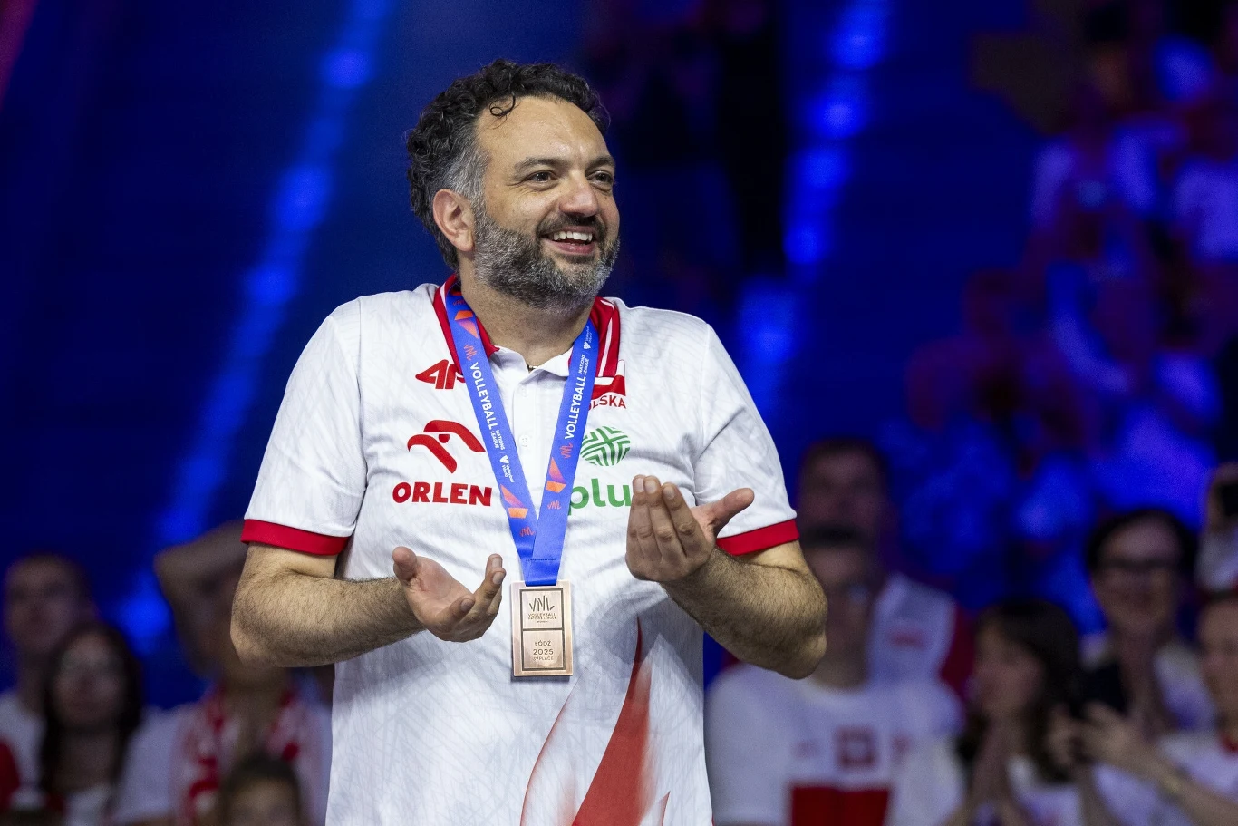 Mężczyzna w białej koszulce z logotypami sponsorów, z medalem na niebieskiej szarfie na szyi, uśmiecha się i gestykuluje rękami, w tle kibice w biało-czerwonych barwach.