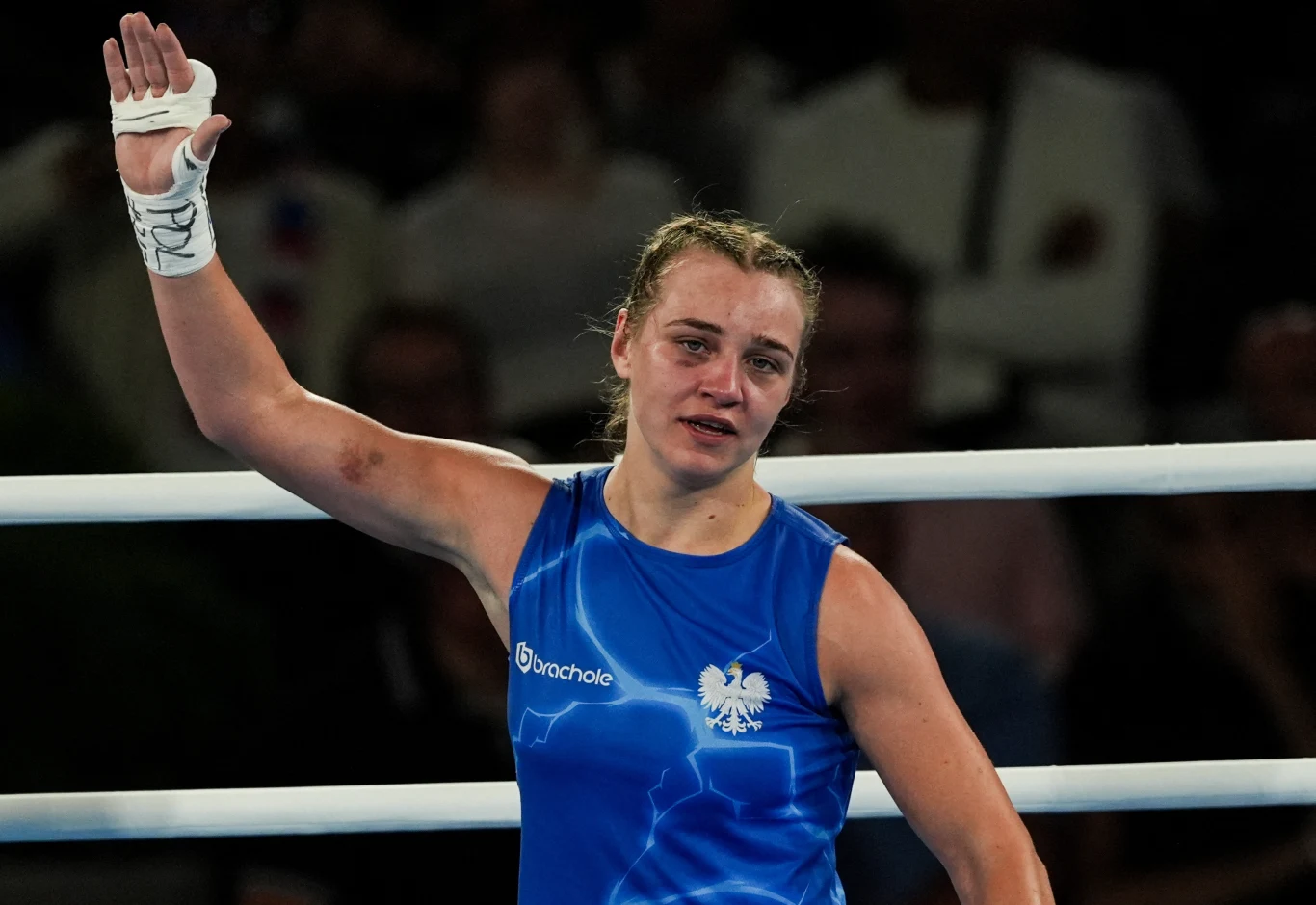 Bokserka w niebieskim stroju z polskim godłem na piersi podnosi rękę w geście zwycięstwa, na twarzy widoczne emocje i ślady zmęczenia, w tle rozmyte sylwetki widzów.