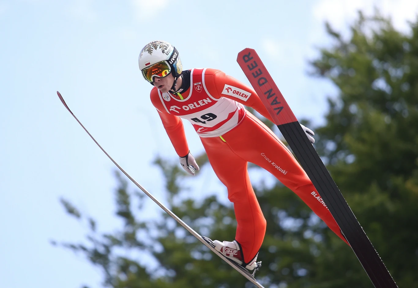 Adam Niżnik Skoczek narciarski w czerwonym kombinezonie i kasku w locie nad śnieżnym podłożem, dynamiczna pozycja na tle błękitnego nieba i zielonych drzew, widoczne profesjonalne narty z napisem VAN DEER.