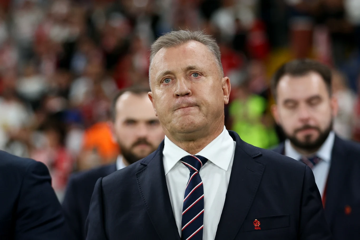 Mężczyzna w garniturze i krawacie z poważnym wyrazem twarzy, stojący na tle osób ubranych formalnie, w otoczeniu stadionu pełnego kibiców.