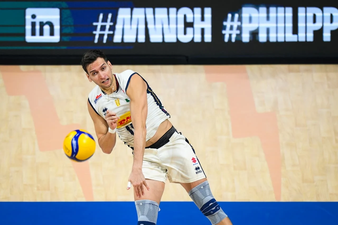 Yuri Romano w trakcie meczu Włochy - Argentyna Siatkarz w stroju sportowym odbijający piłkę podczas meczu halowego, dynamiczna pozycja oraz wyraźne skupienie na grze; na tle widoczne oznaczenia i hashtagi na bandach reklamowych.