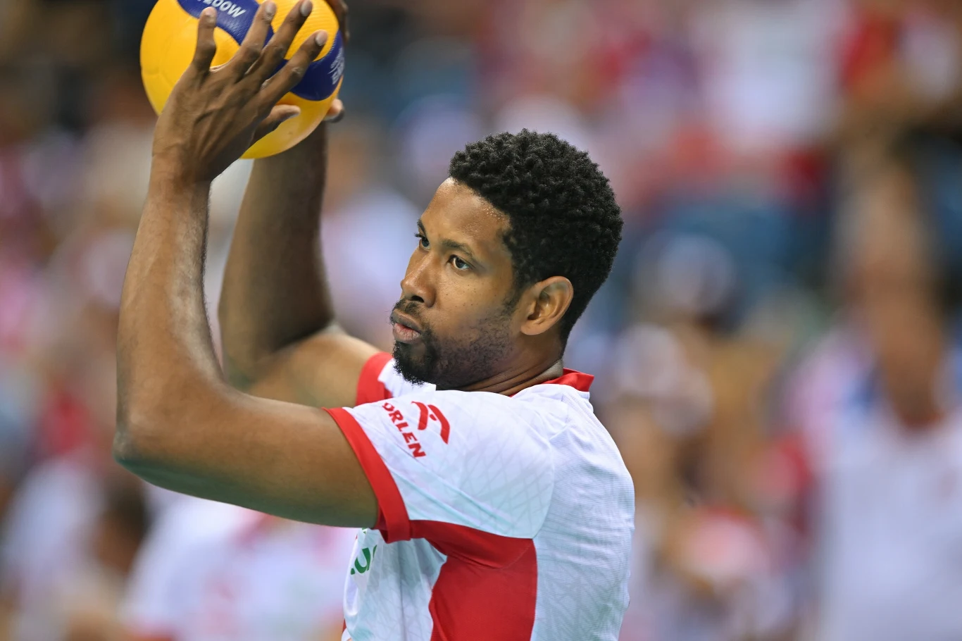 Wilfredo Leon Siatkarz trzymający piłkę do siatkówki w gotowości do rozegrania akcji, ubrany w biało-czerwony strój sportowy, w tle widoczna rozmyta publiczność.