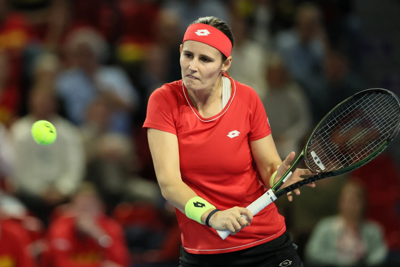Tenisistka w czerwonej koszulce oraz opasce na głowie koncentruje się na odbiciu piłki tenisowej podczas meczu, trzymając rakietę obiema rękami; w tle rozmazany tłum kibiców.